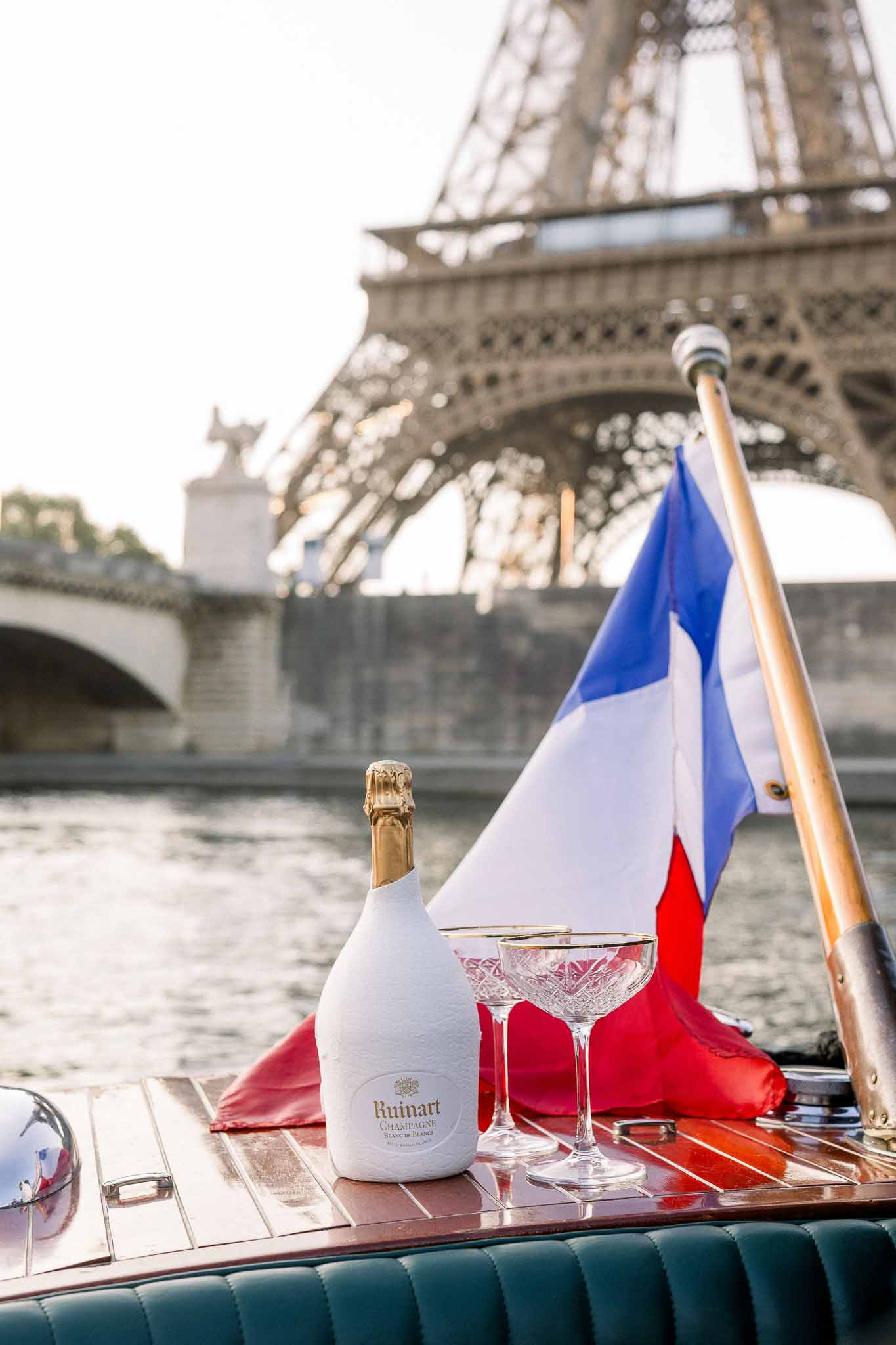 Champagne bottle and gold-rimmed coupe glasses on wooden boat deck with Eiffel Tower in background