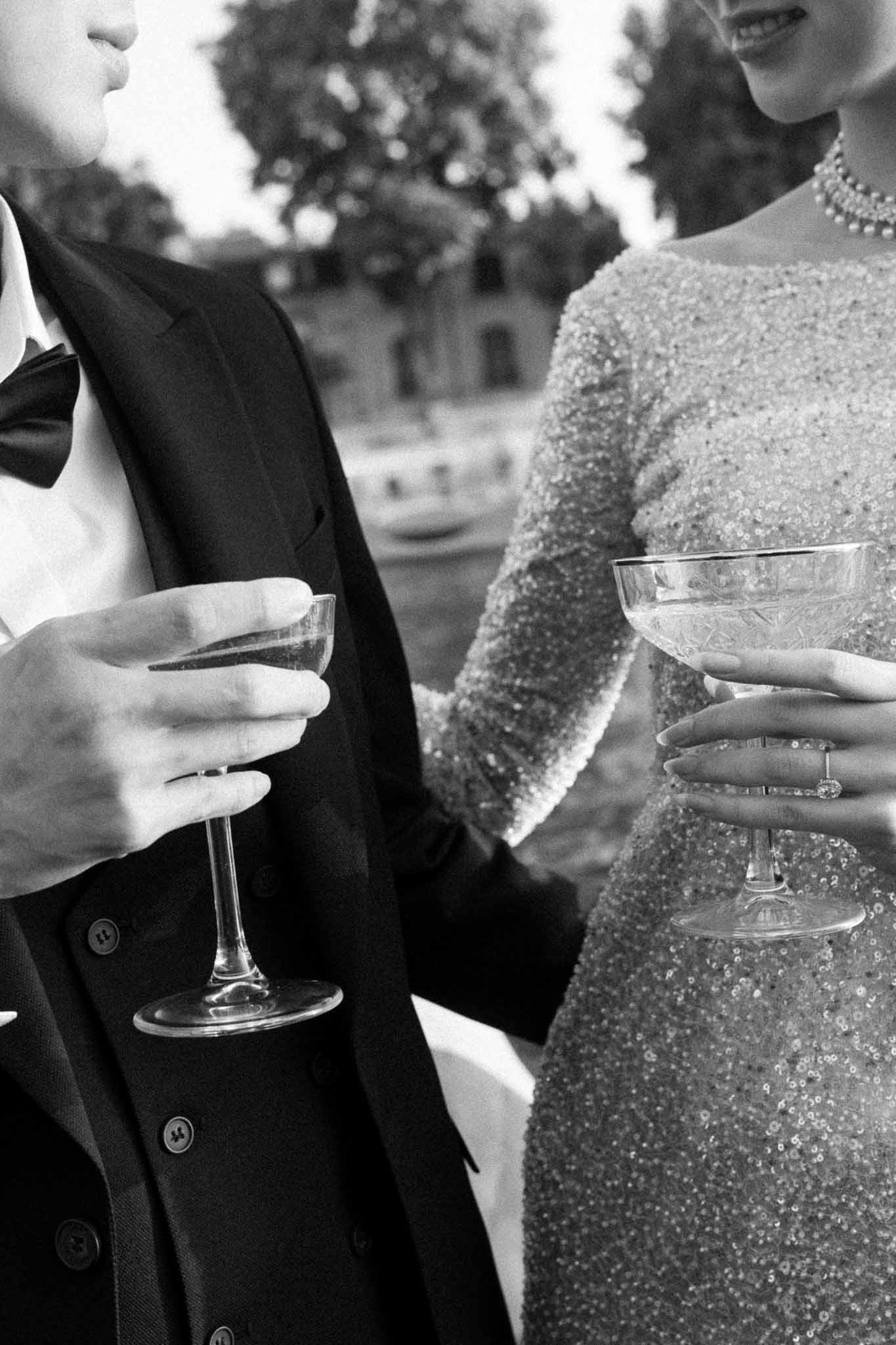 Close-up of bride in beaded dress and groom in tuxedo holding champagne coupes with diamond ring visible in B&W