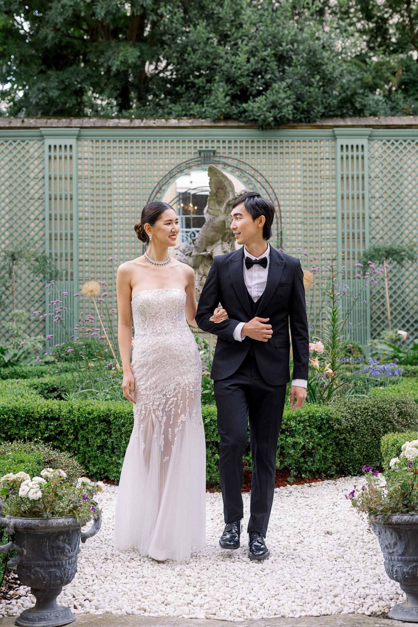 Bride in beaded gown and groom in black tuxedo walking arm-in-arm through formal garden with rose urns