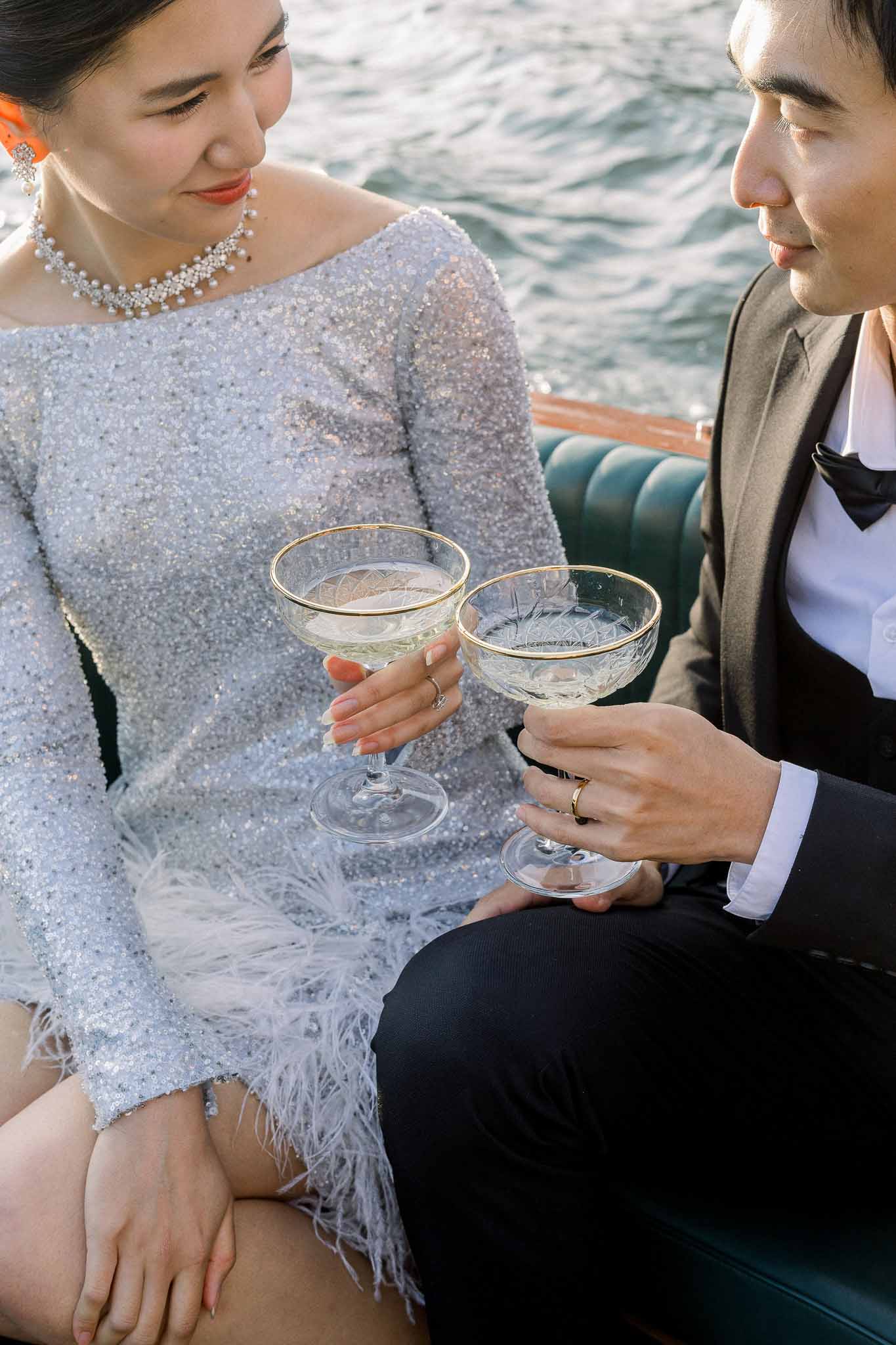 Couple toasting champagne in gold-rimmed coupe glasses on boat with bride in silver sequin mini dress