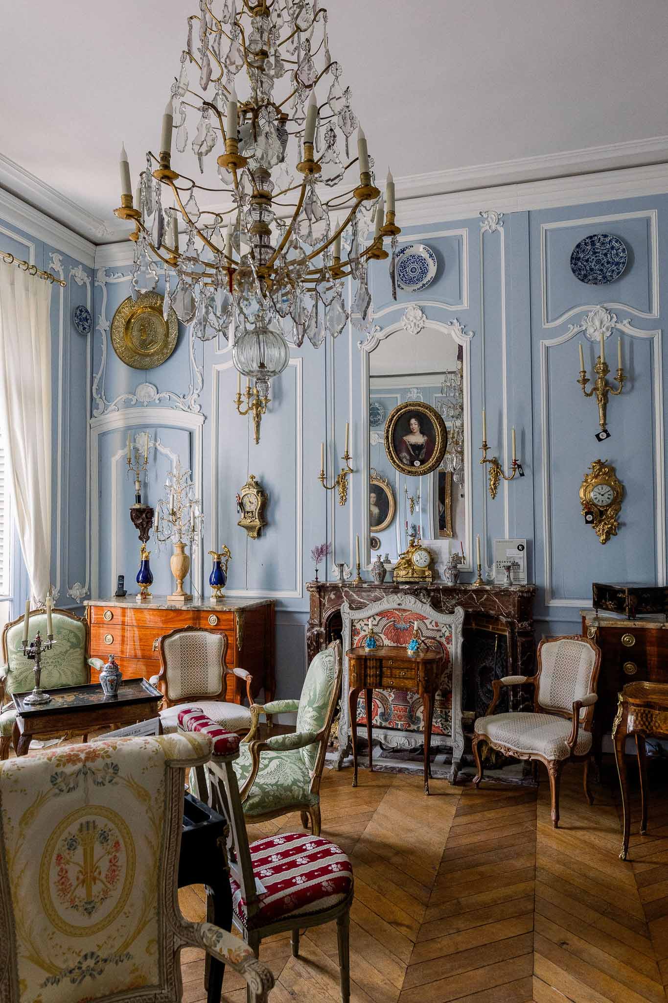 Powder-blue paneled salon with crystal chandelier, gilt fireplace, decorative plates, and Louis XV armchairs