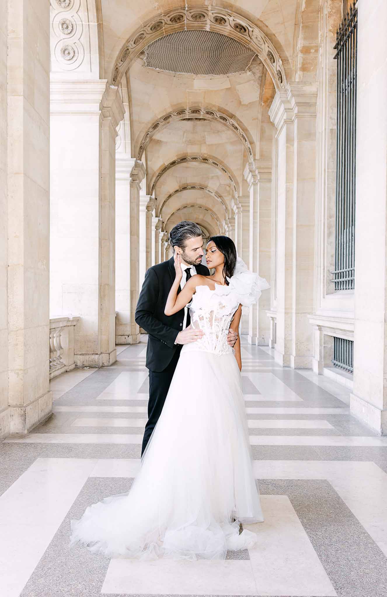 Bride in ruffled organza bodice and groom in black suit touching foreheads in Parisian colonnade gallery