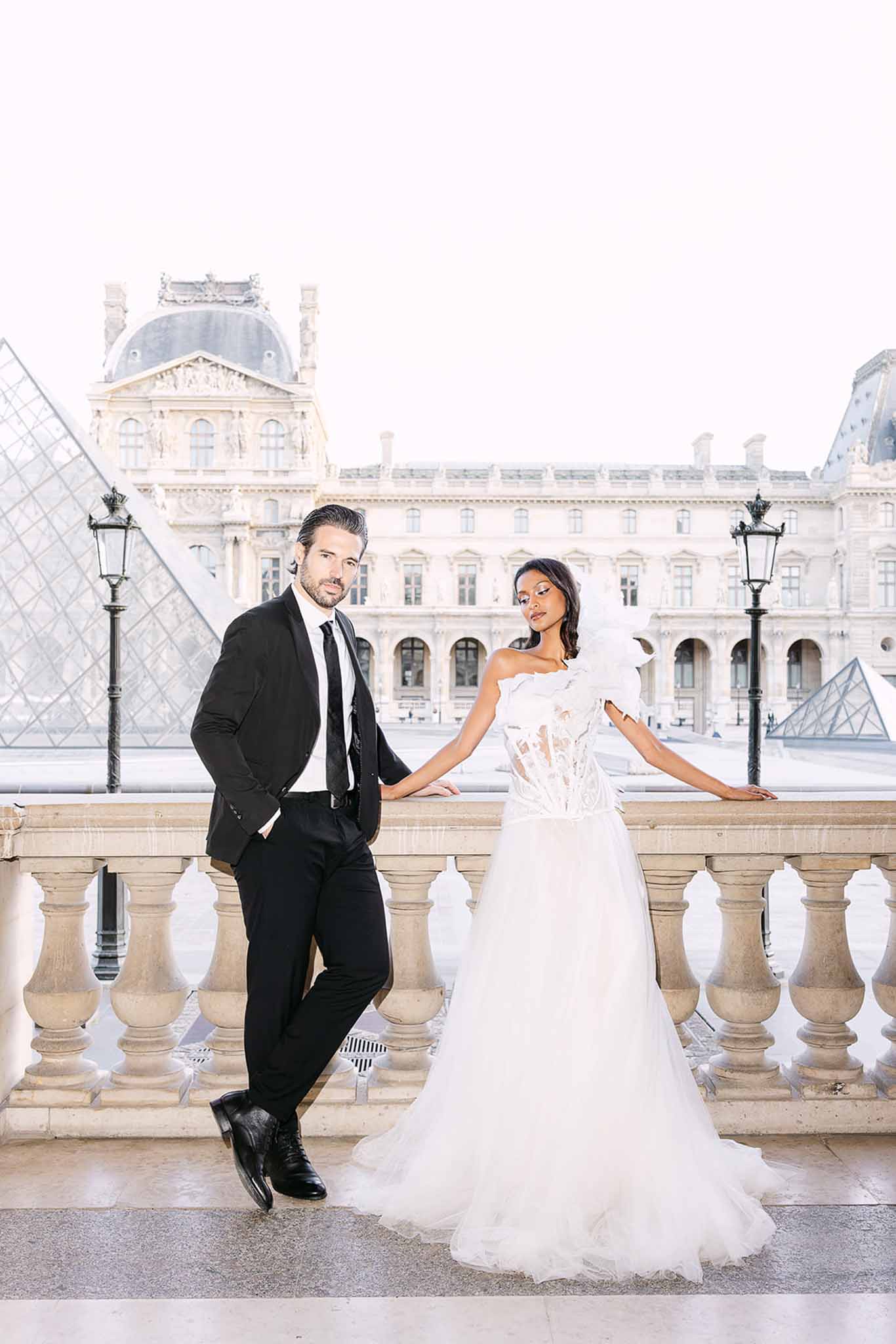 Bride in one-shoulder tulle gown and groom in black suit pose on Louvre terrace with glass pyramid behind