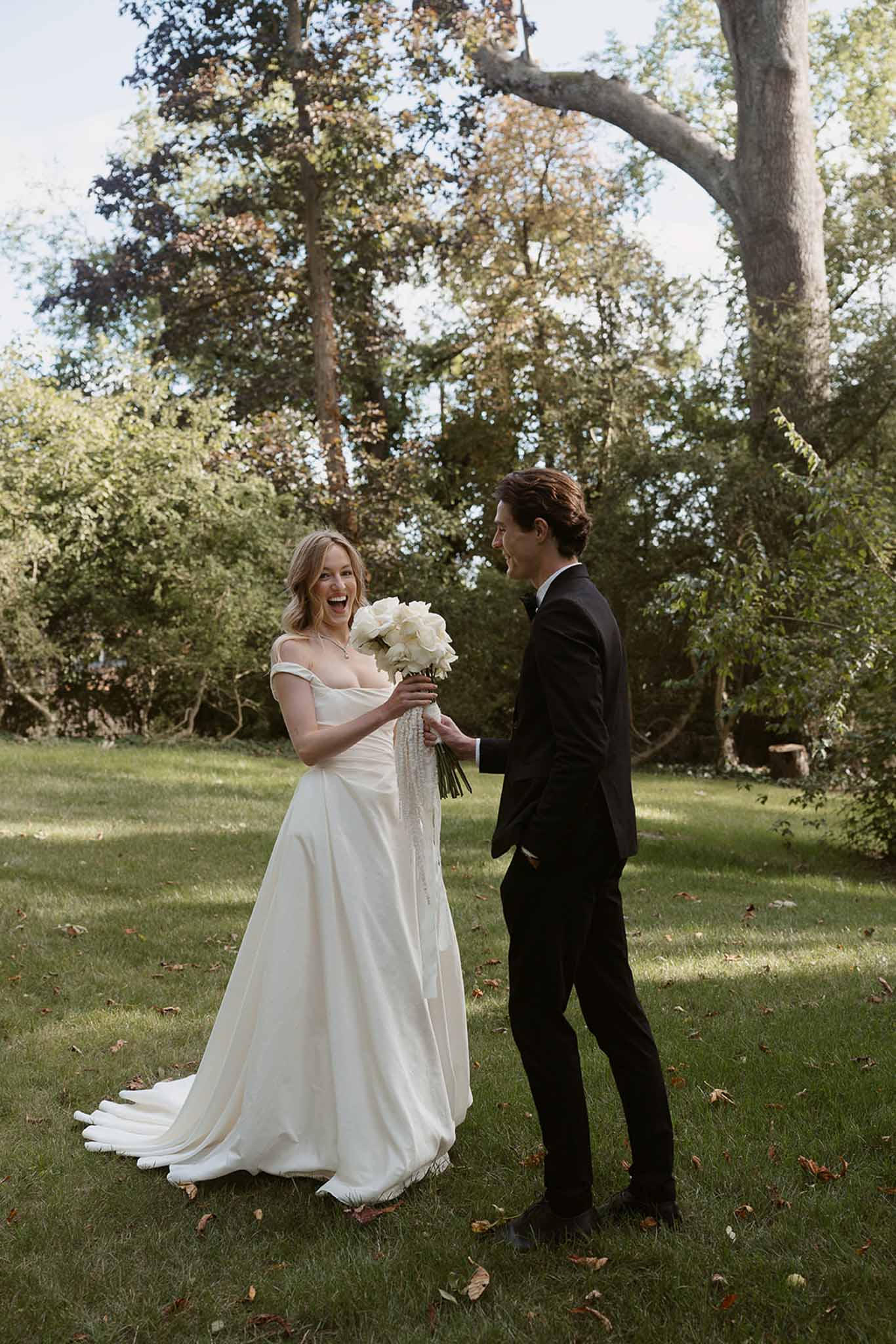Couple laughing on garden lawn as groom playfully reaches for bride's white peony bouquet