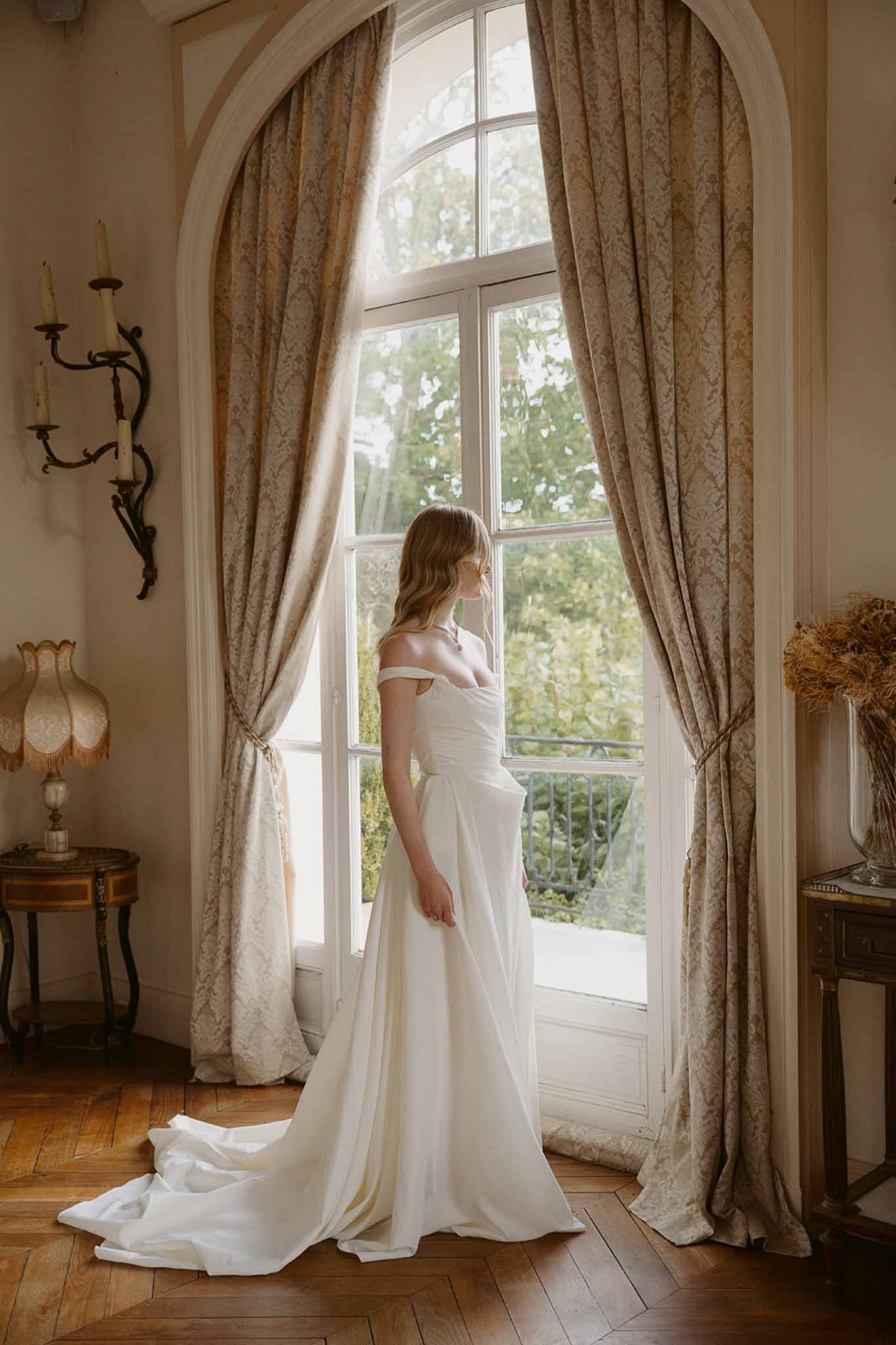 Bride in off-the-shoulder gown standing at arched window inside a French chateau interior