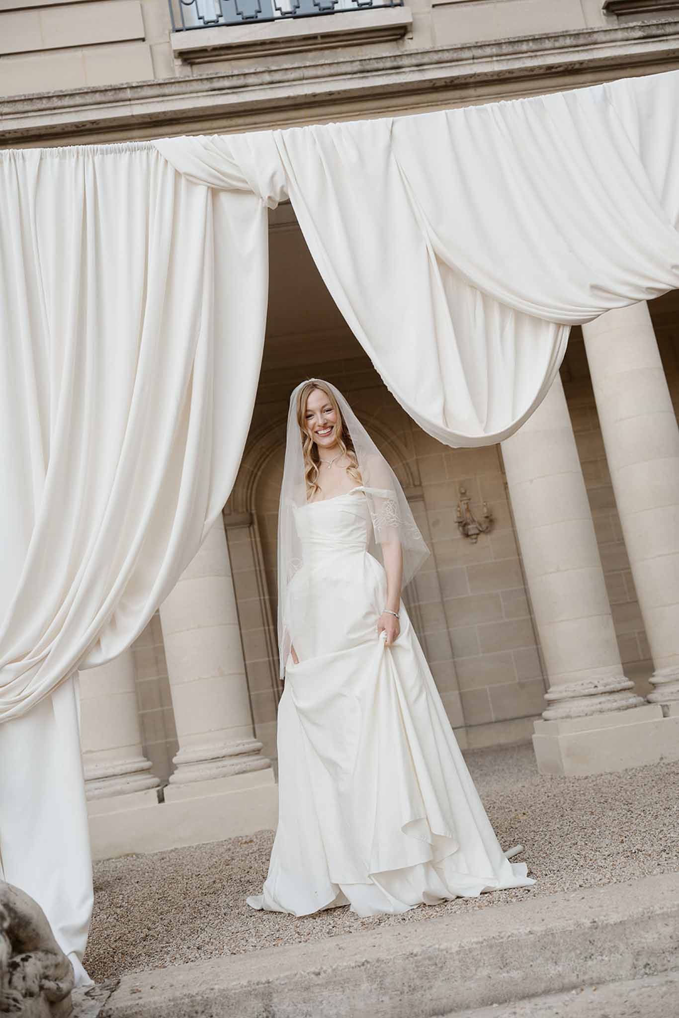 Bride in ivory strapless satin gown with cathedral veil beneath draped fabric canopy at limestone chateau columns