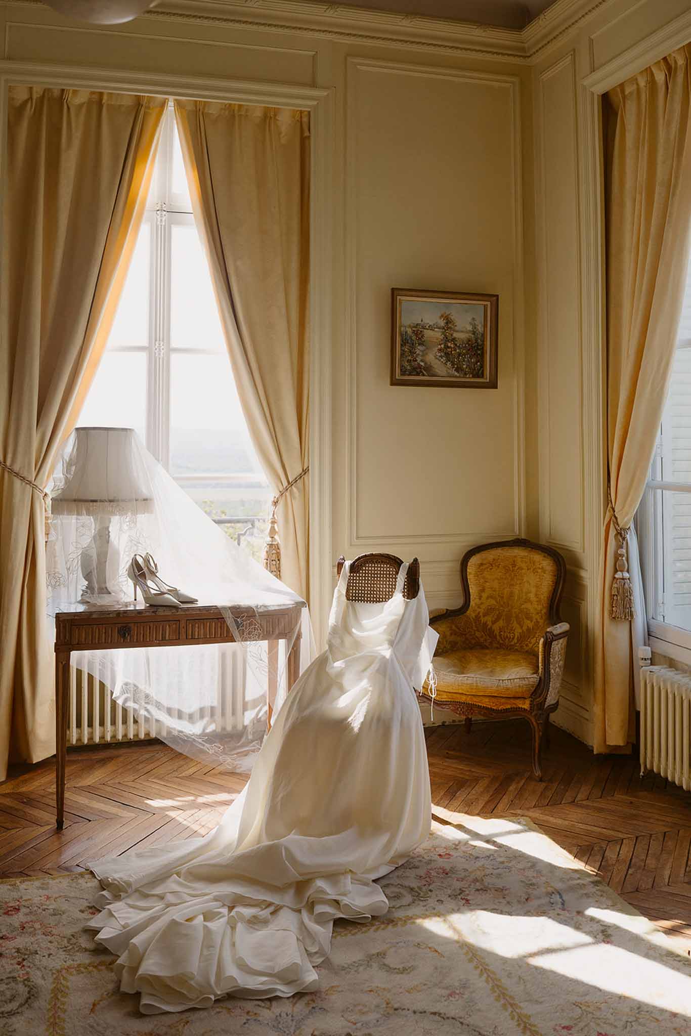 Wedding dress and lace veil draped over cane-back chair with bridal shoes on console table in chateau bridal suite