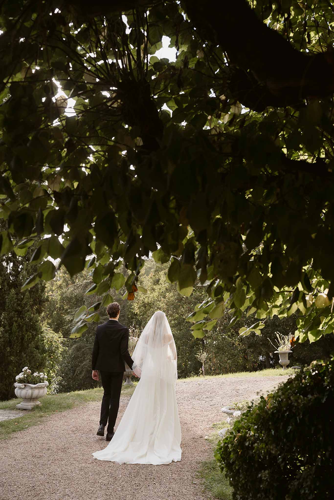 Couple walking hand in hand away from camera with cathedral veil trailing on formal garden path