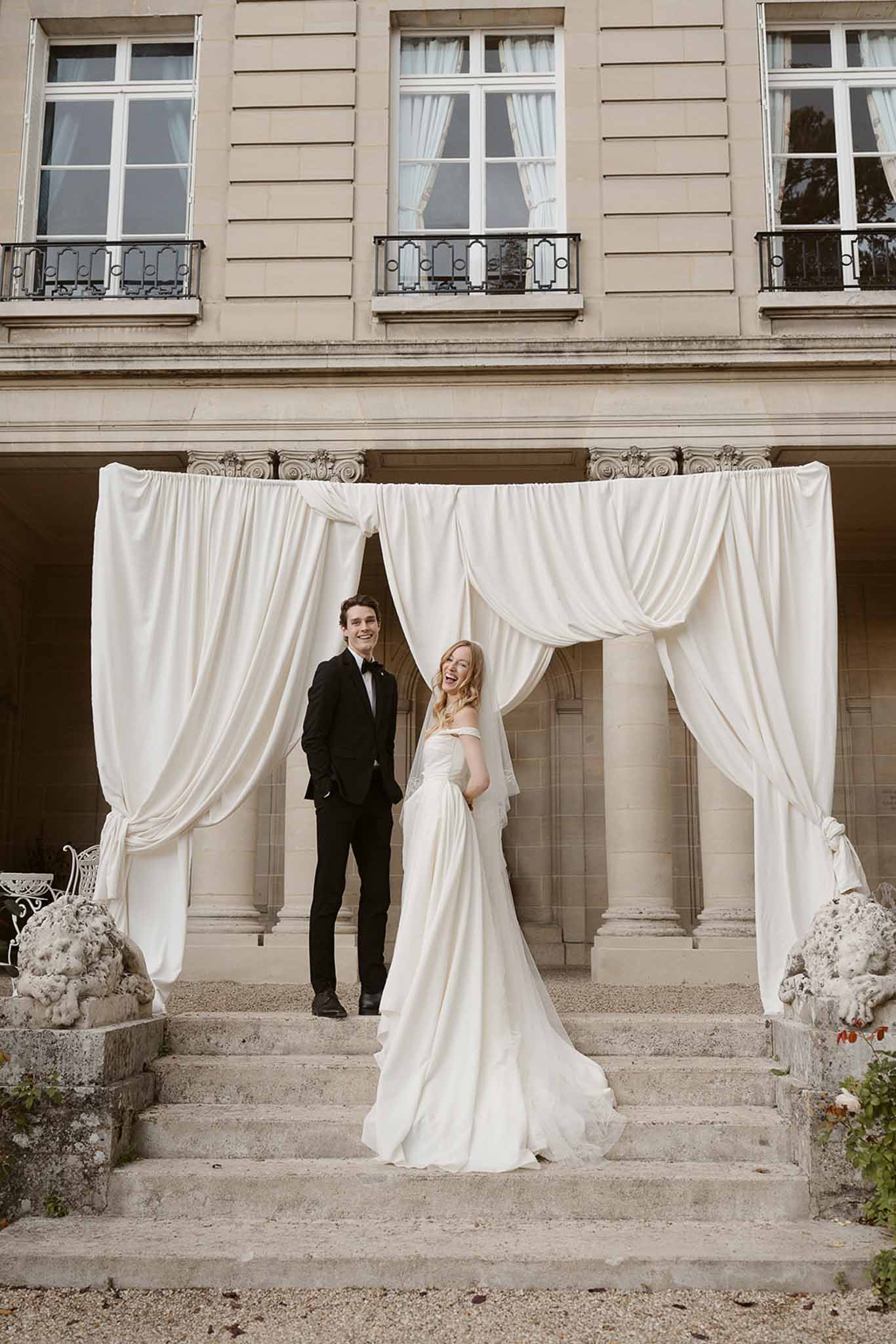 Laughing couple on chateau steps with ivory draped arch, bride in cathedral veil and off-shoulder gown