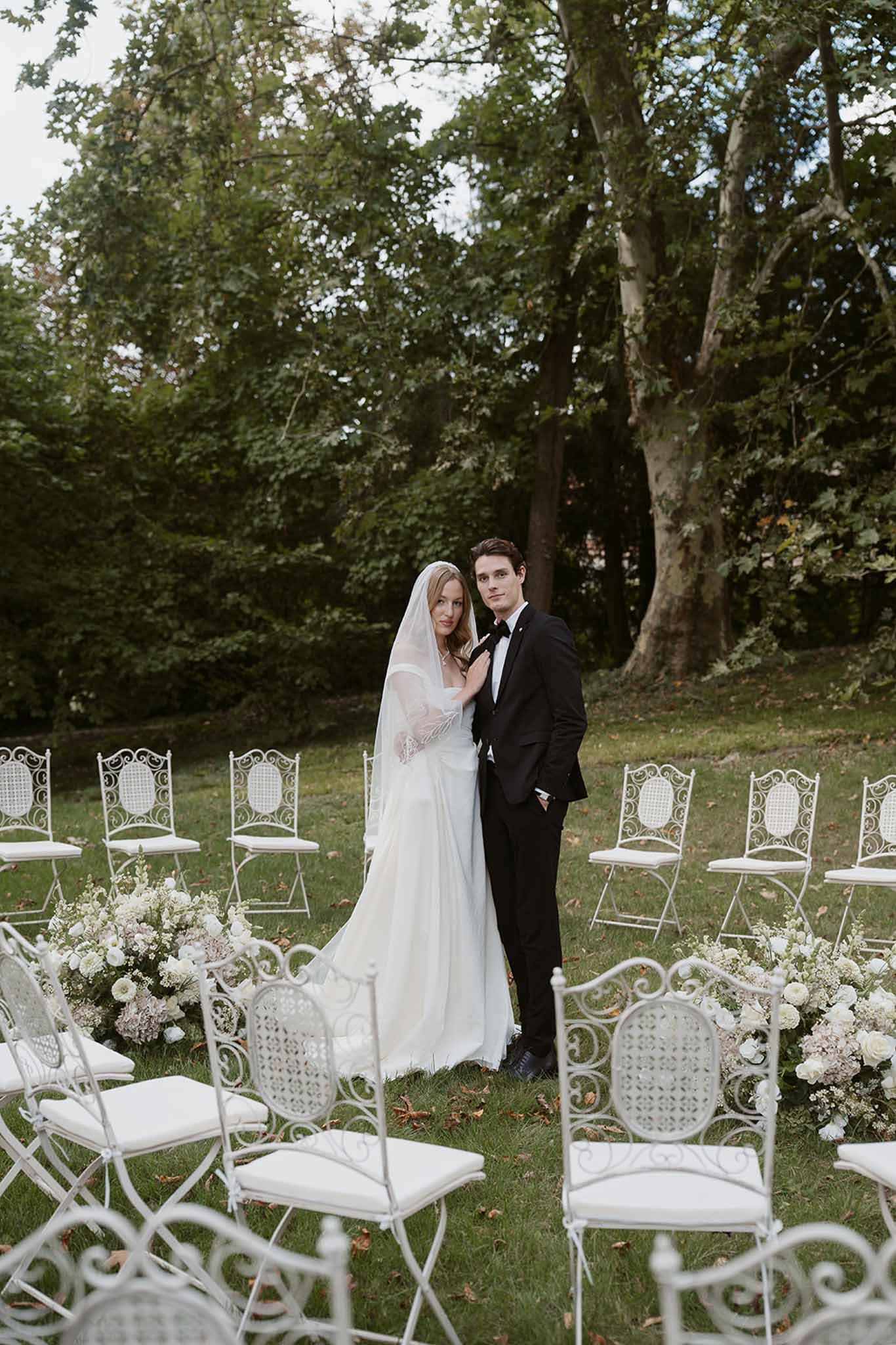 Bride and groom at outdoor garden altar flanked by white and blush floral arrangements with wrought-iron chairs