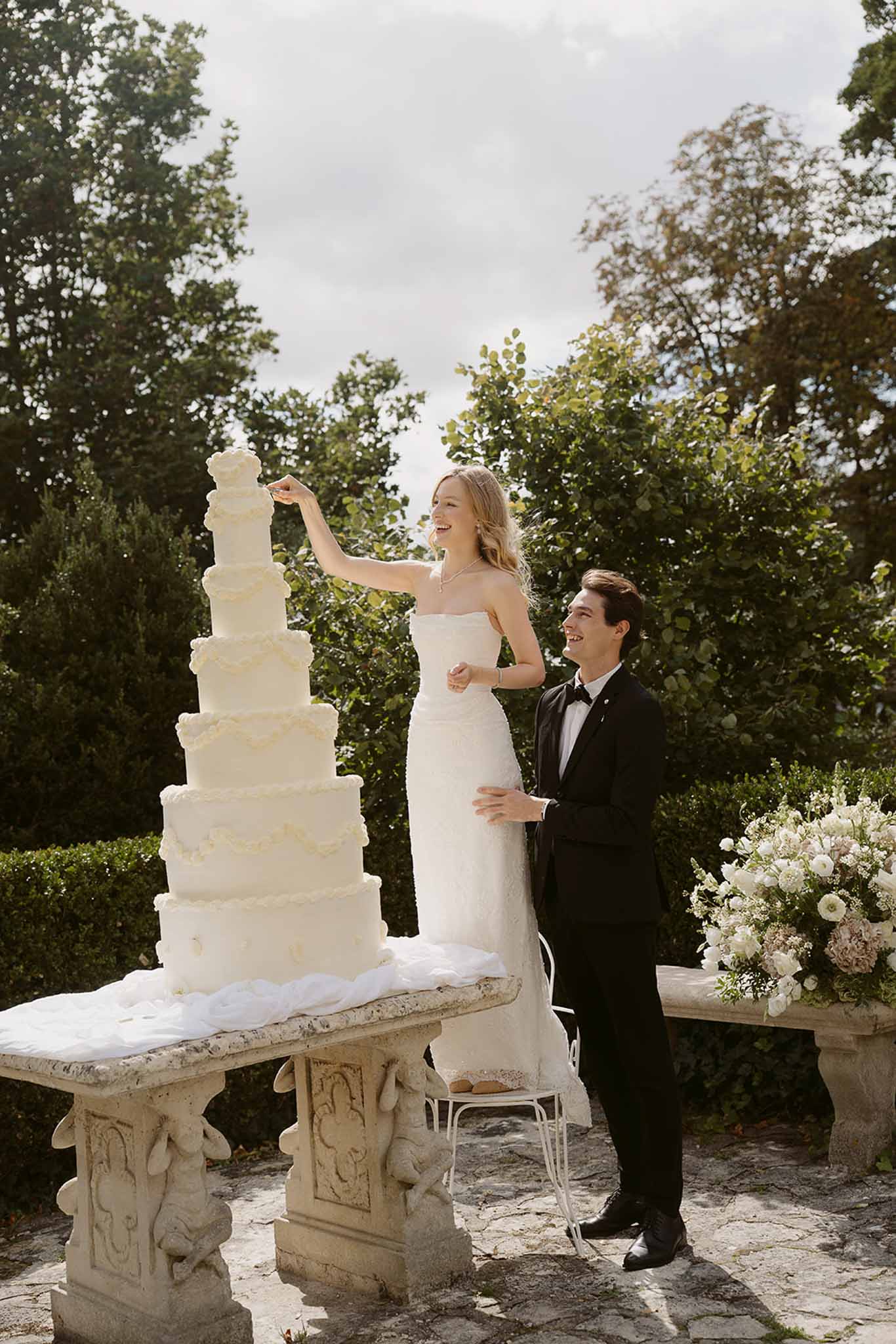 Bride and groom laughing beside six-tier white wedding cake on stone pedestal outdoors
