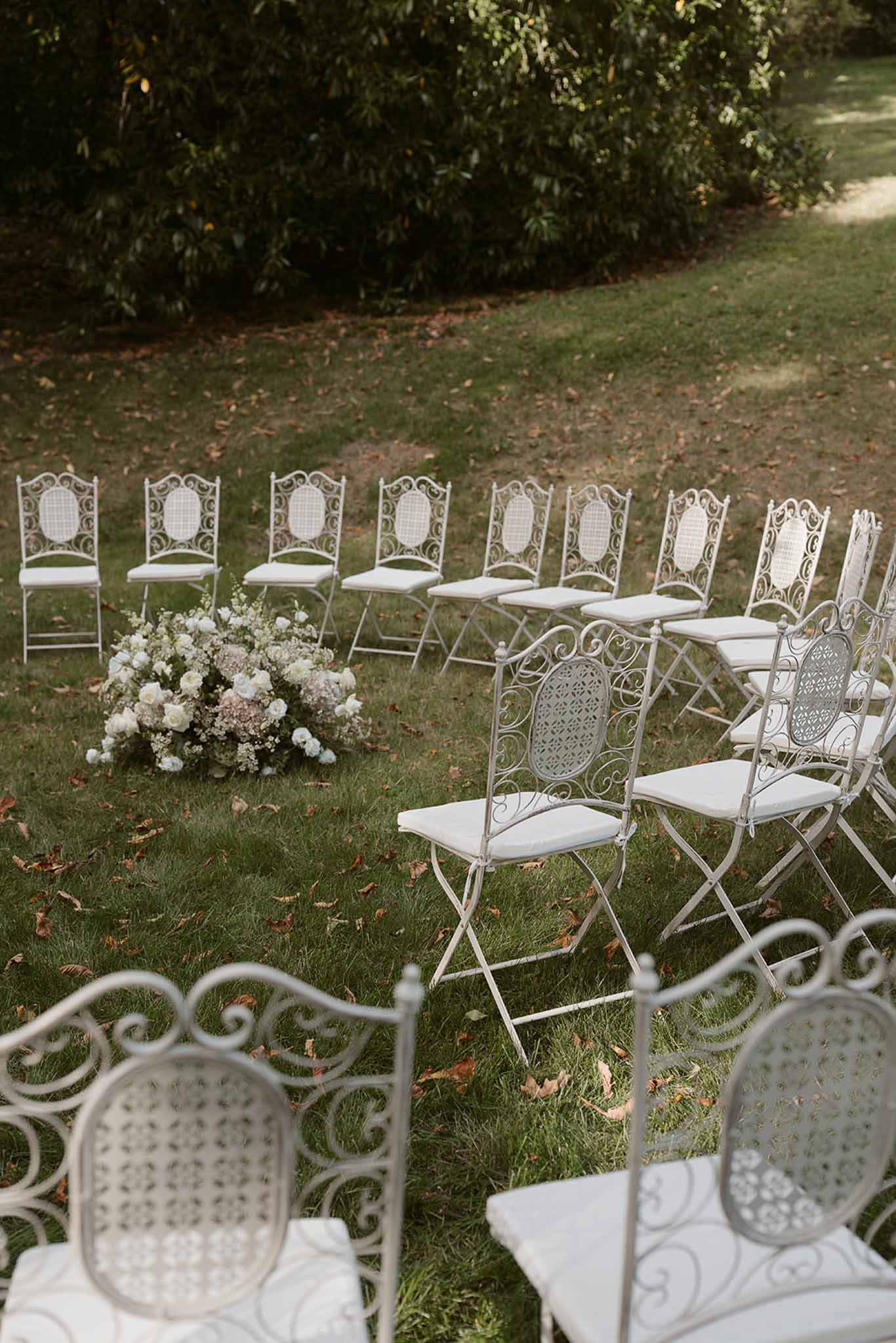Empty ceremony setup with white wrought iron chairs on lawn and ground-level white rose and mauve hydrangea cluster