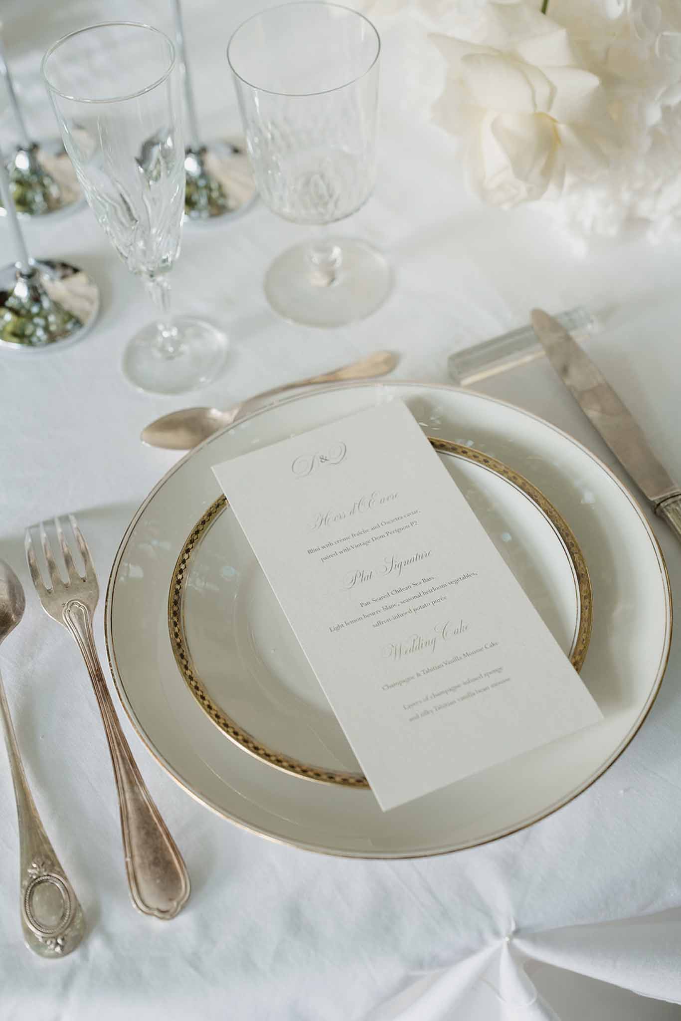 Reception place setting with gold-rimmed charger plates, printed menu card, crystal glassware, and white peonies