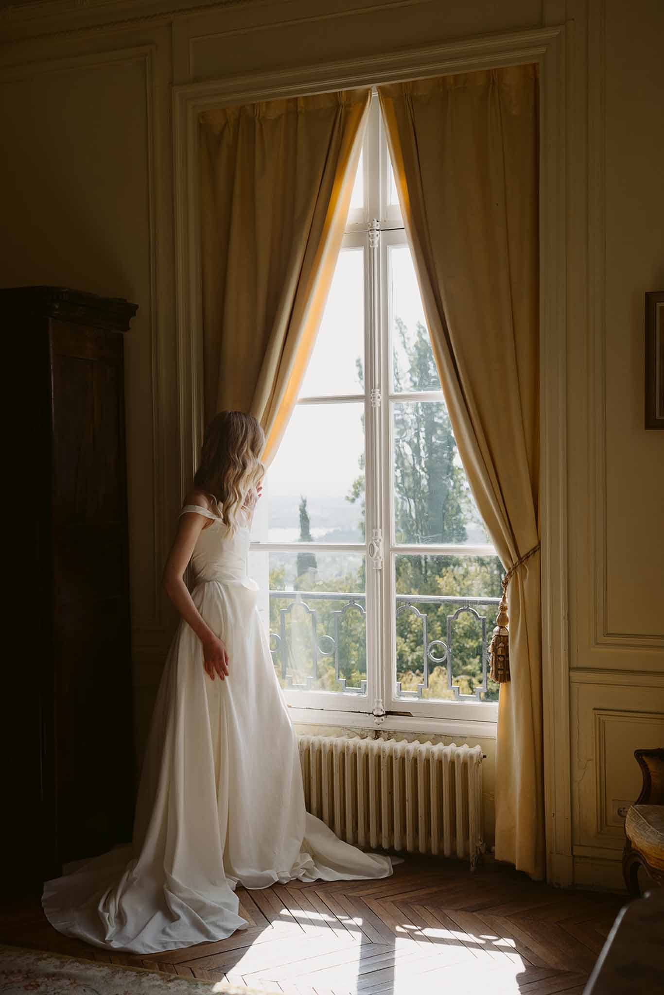 Bride in off-the-shoulder ivory gown standing at tall chateau window looking out, photographed from behind