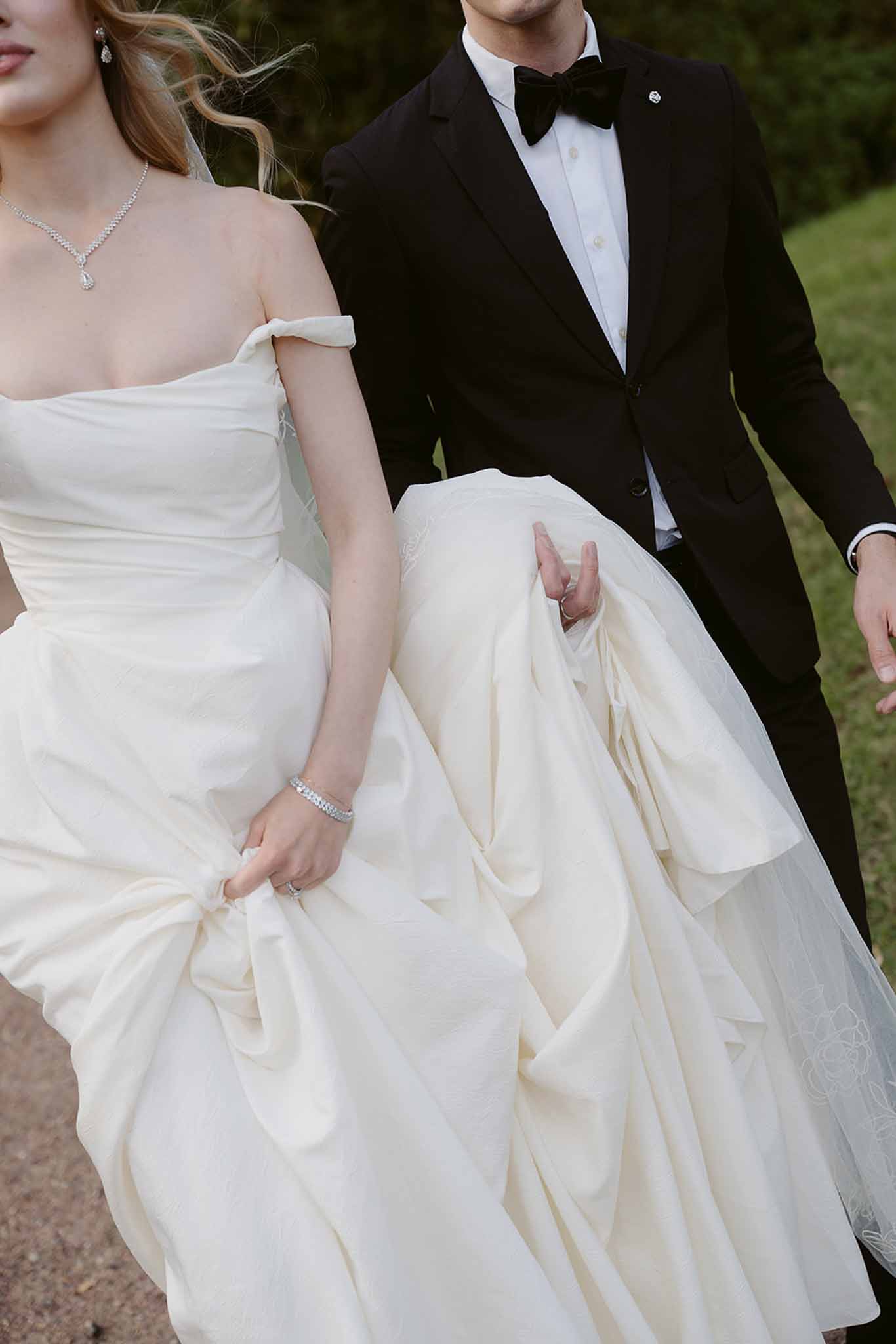 Torso-level close-up of couple walking, bride gathering embroidered ballgown with diamond bracelet and pendant