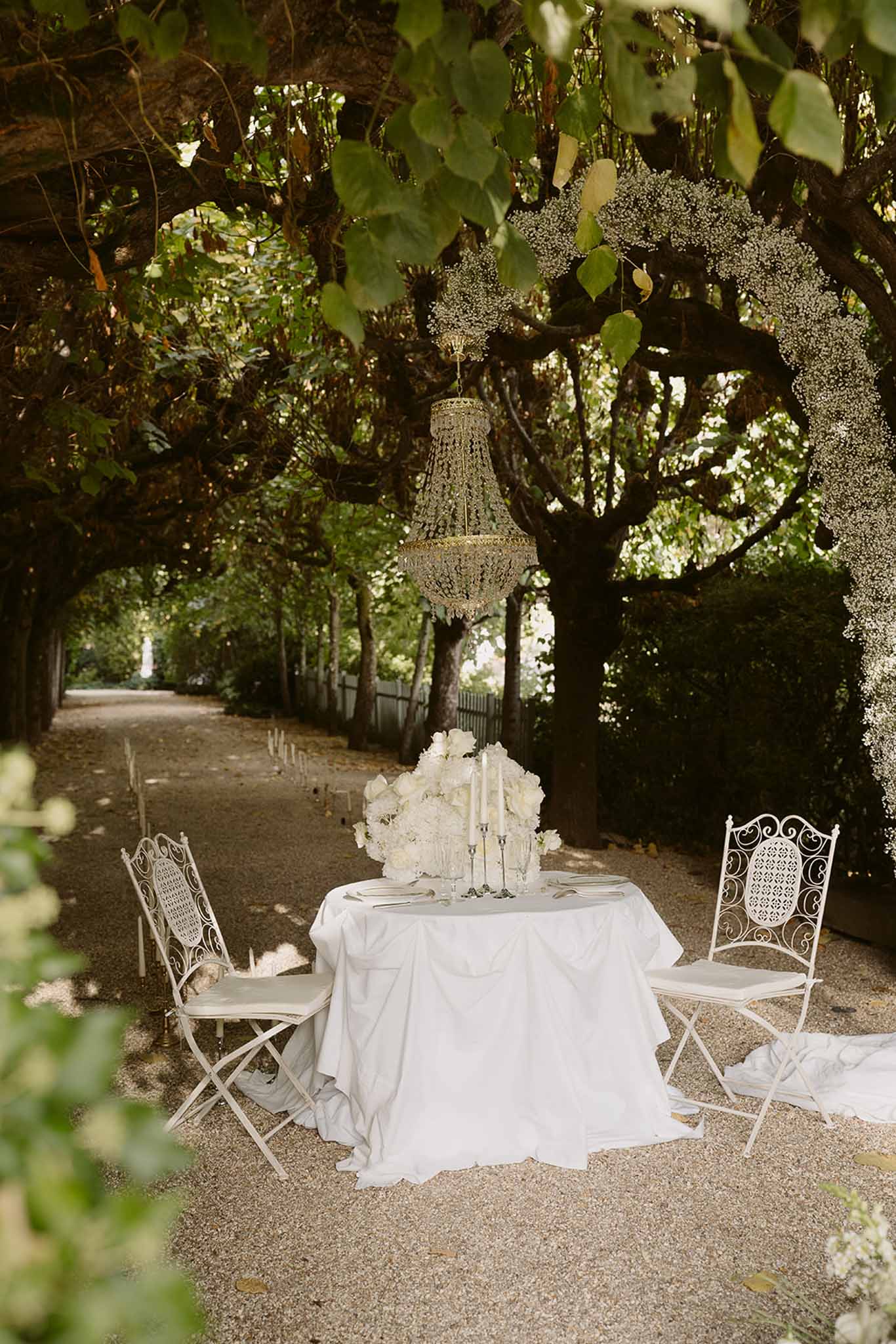 White sweetheart table under gold chandelier in tree-lined allee with roses, peonies, and taper candles