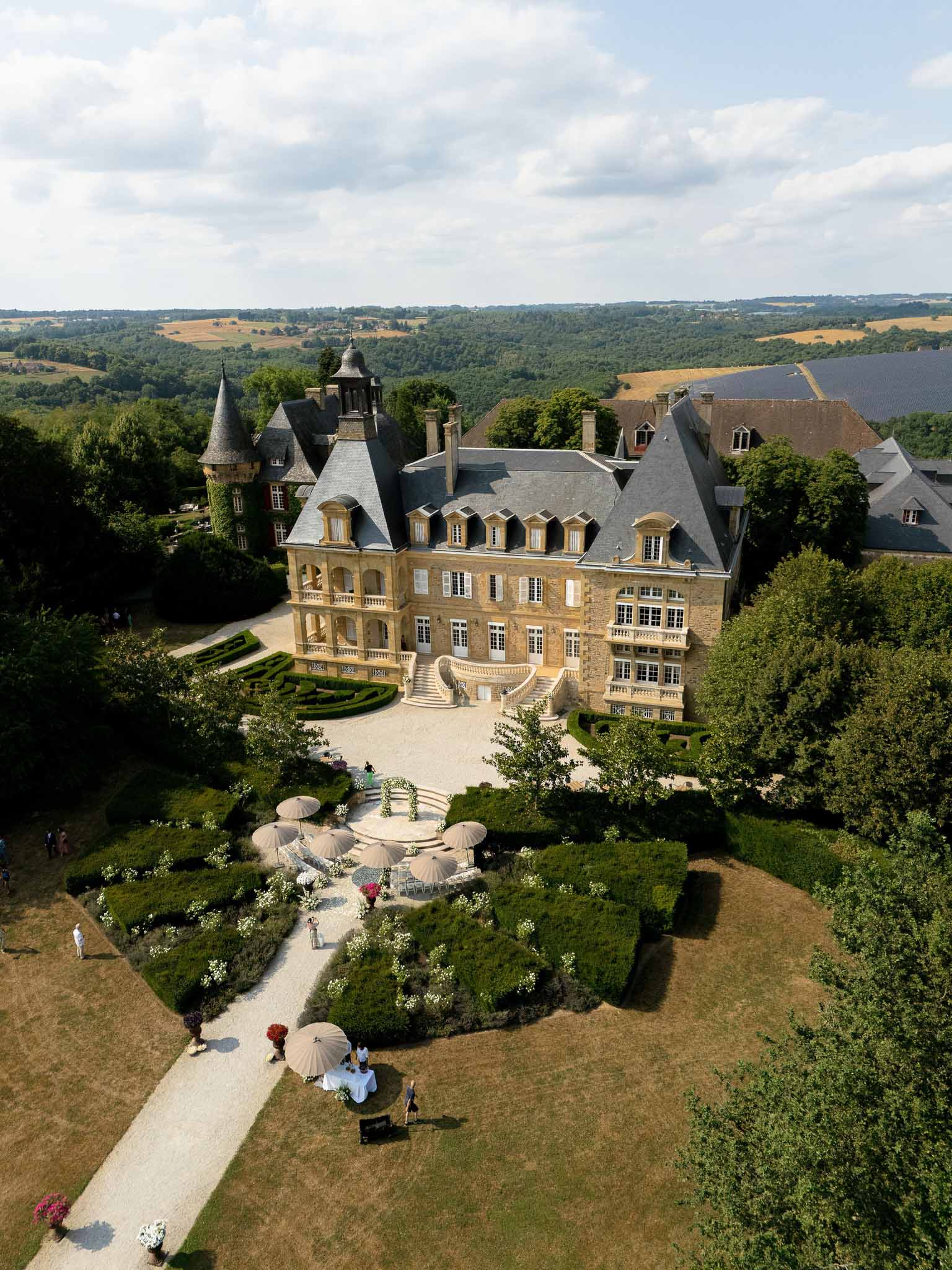 Aerial view of chateau cocktail hour with umbrellas, white tables, and floral arch by grand staircase