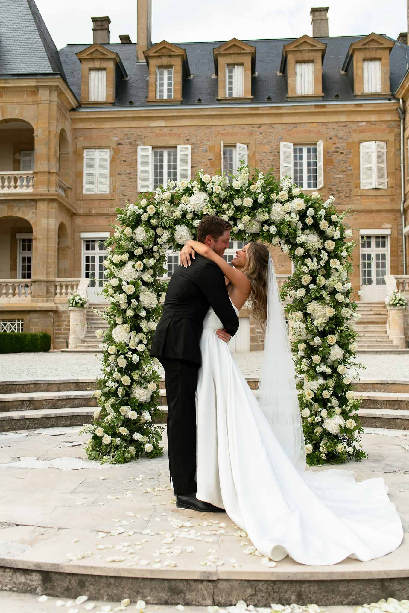 First kiss under circular ivory rose and hydrangea arch on stone terrace before limestone chateau