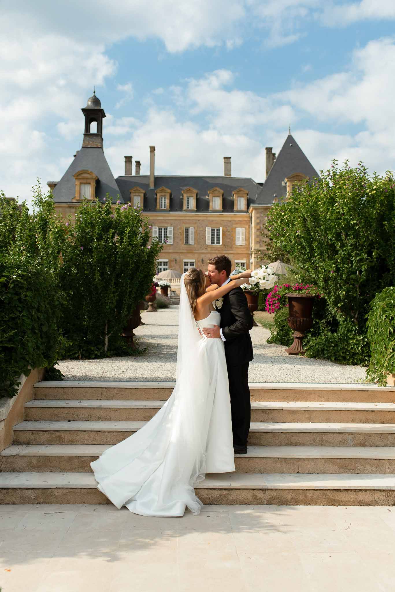 Couple kissing on stone staircase from behind with cathedral train and veil before honey-stone chateau with turret