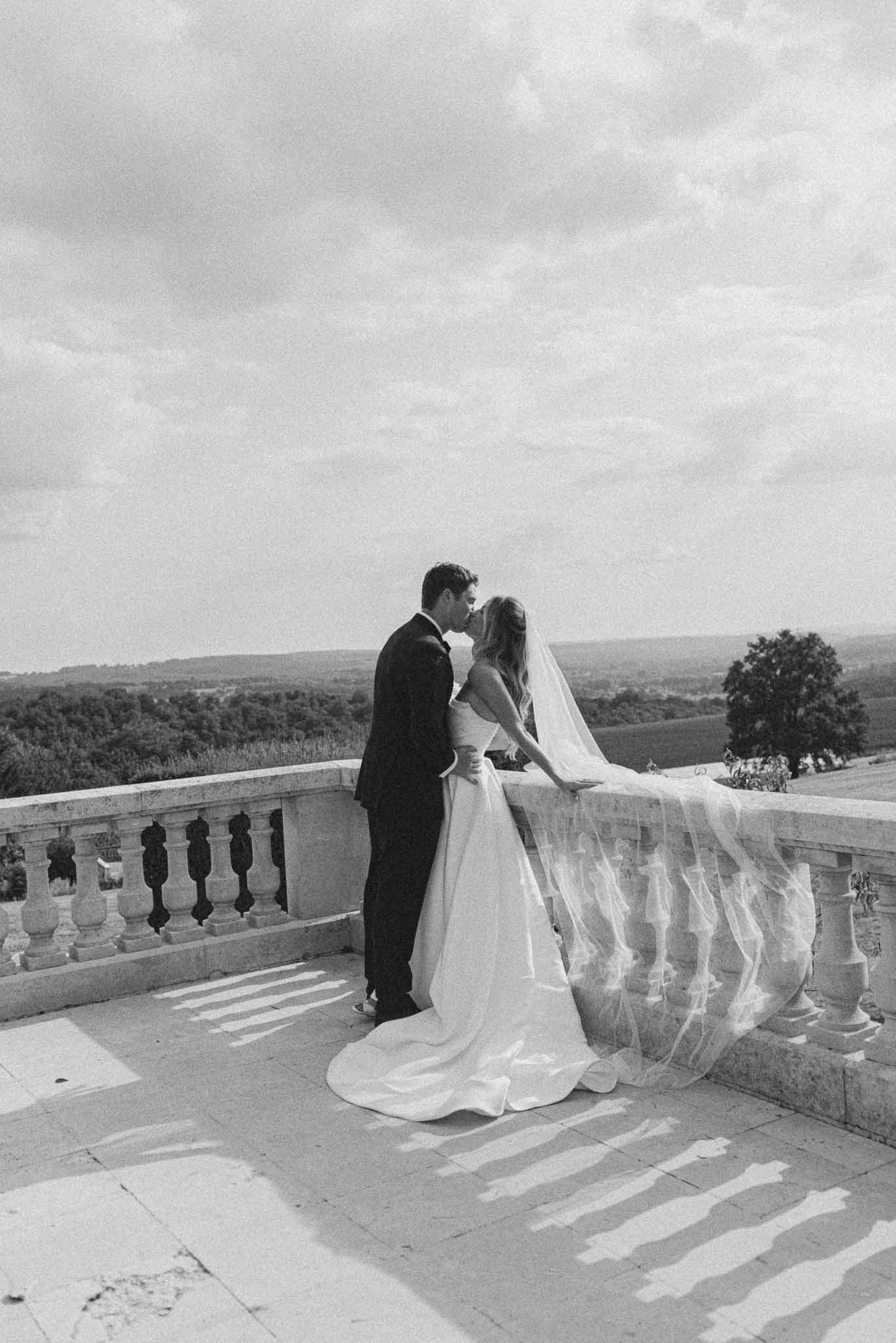 Black and white portrait of bride and groom kissing on stone terrace with cathedral veil and countryside view