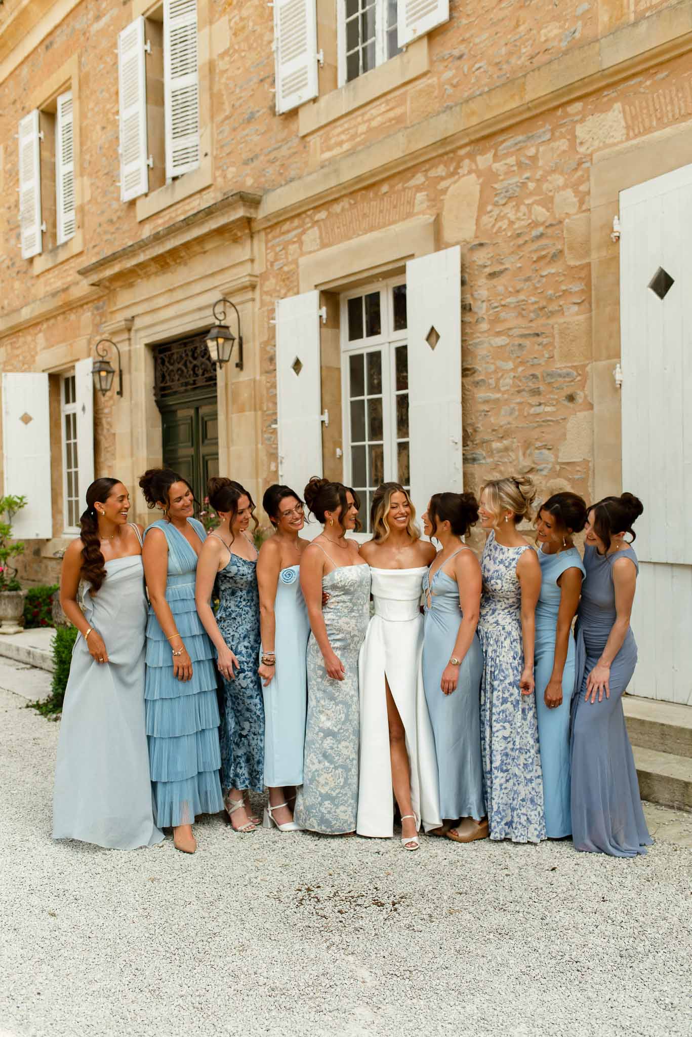 Bride in strapless gown with nine bridesmaids in mix-and-match blue dresses before golden stone chateau