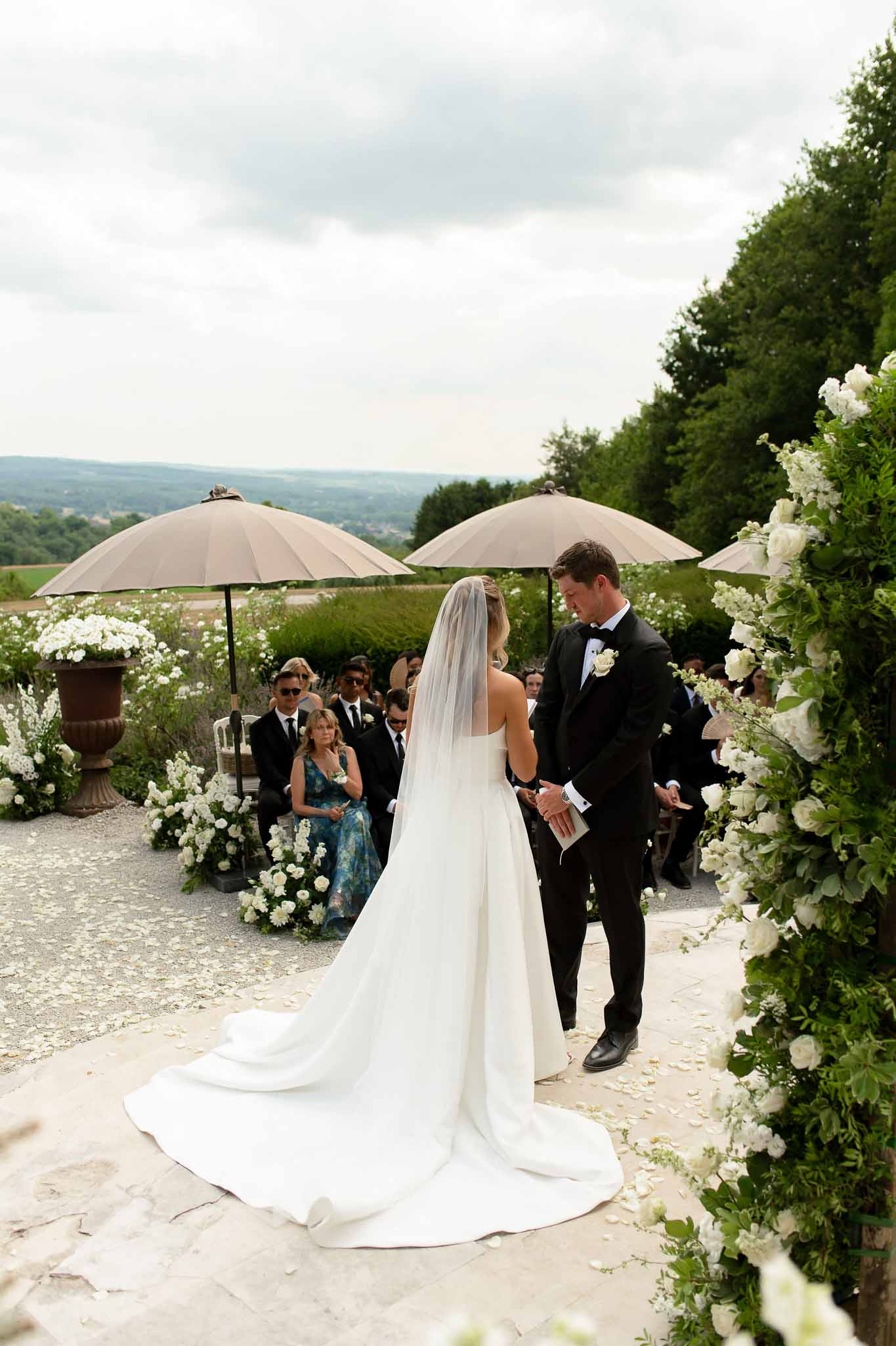 Bride and groom exchanging vows under white rose arch with iron urn arrangements and countryside view