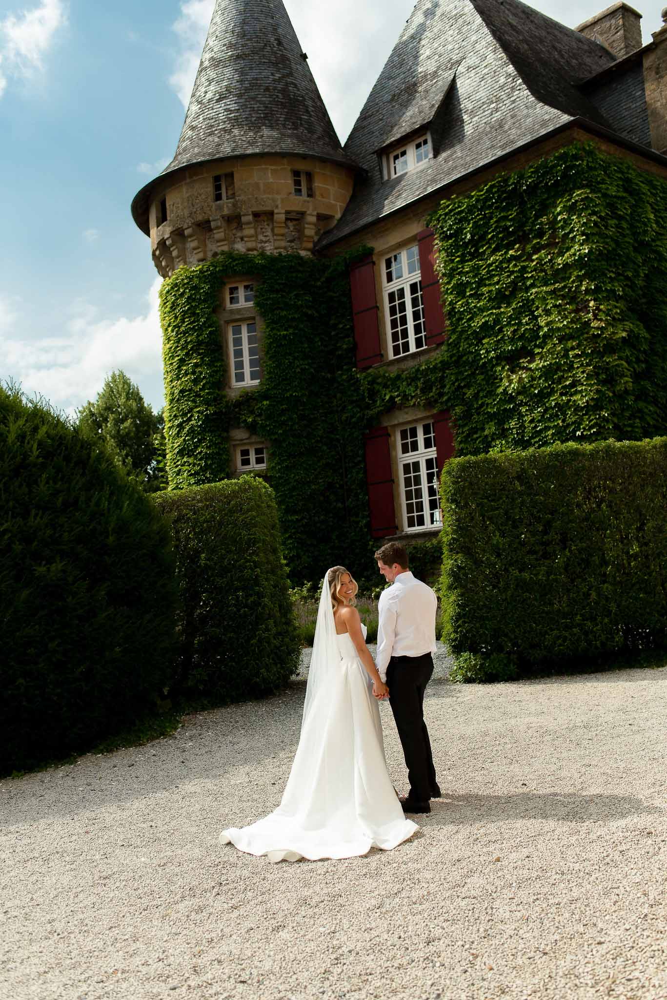 Couple holding hands on gravel before ivy-covered chateau with turrets and red shutters, bride's train trailing