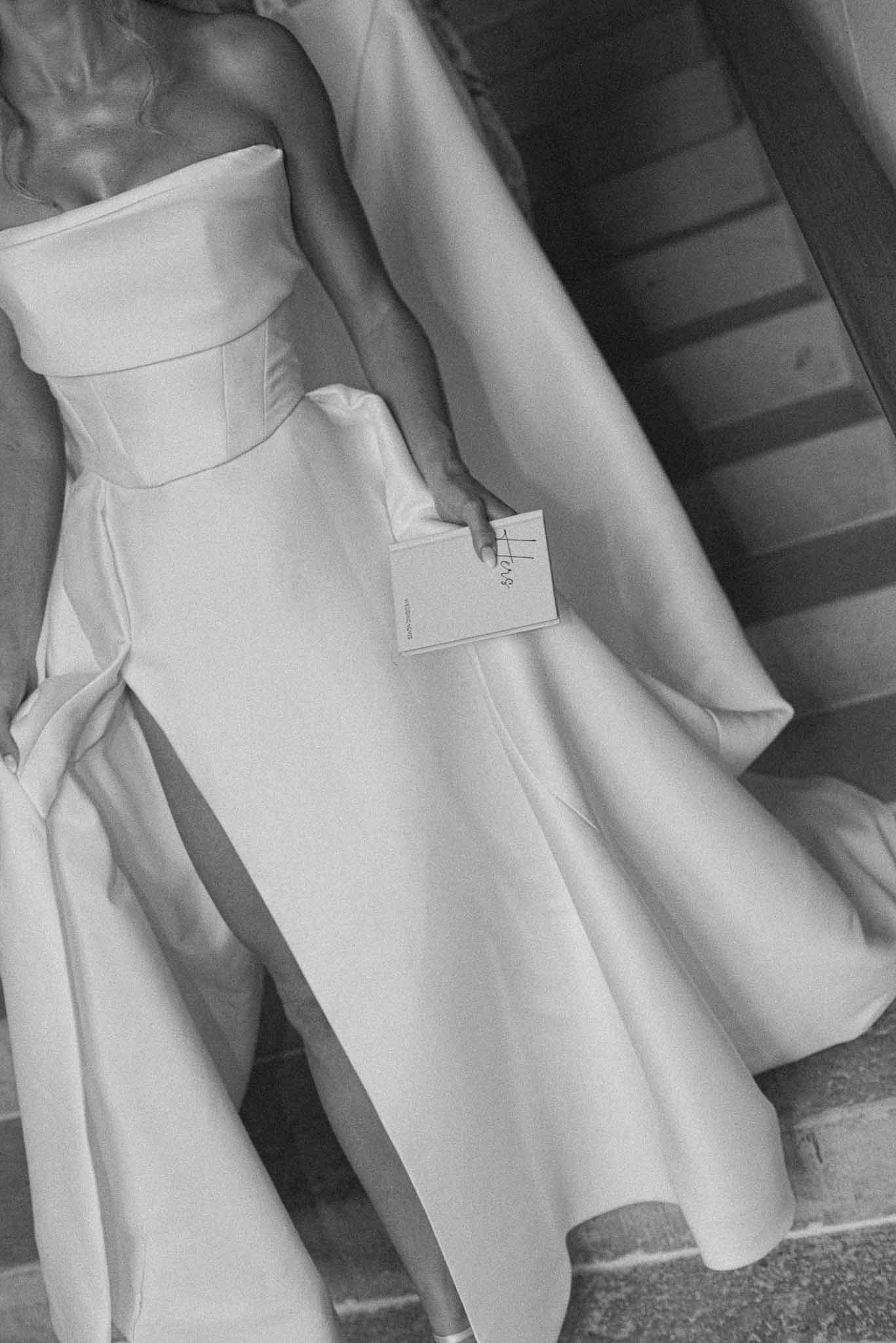 Black and white detail of bride's strapless corset gown holding vow card on stone stairs