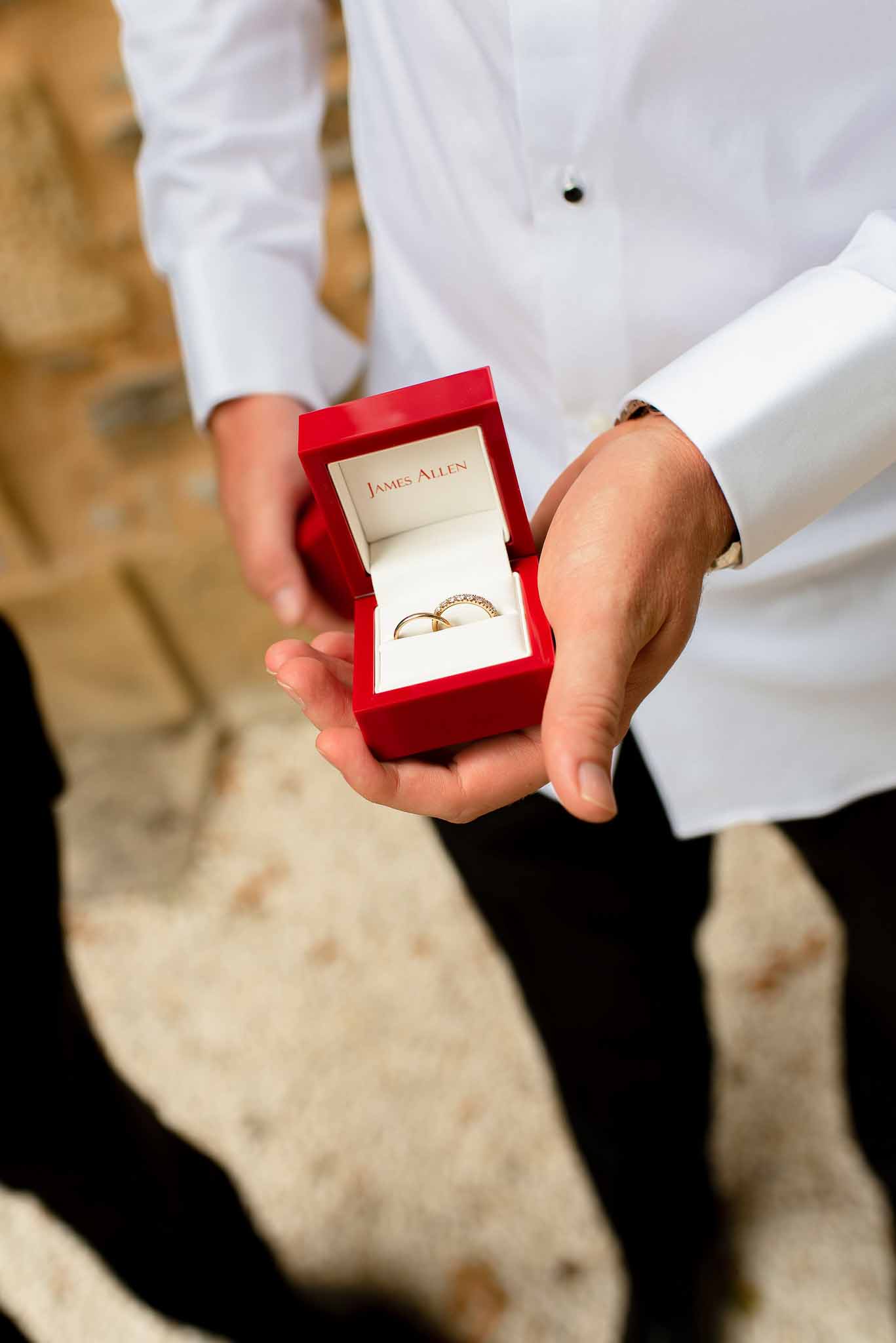 Man holding open red ring box with plain gold band and diamond eternity band outdoors