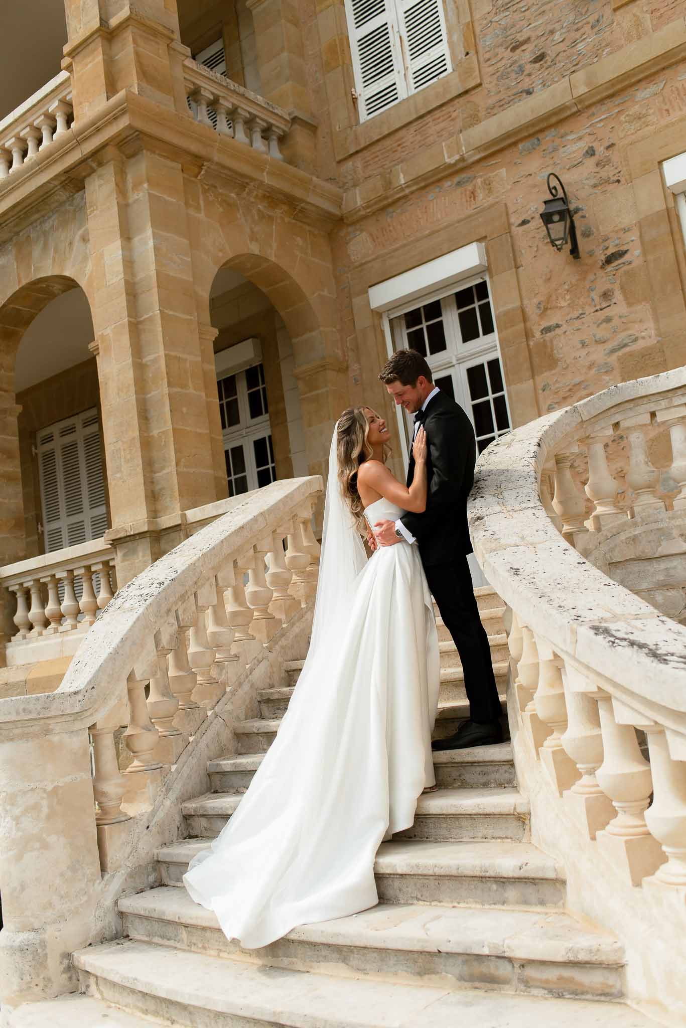 Bride in strapless ball gown and cathedral veil embracing groom in black tuxedo on chateau stone staircase