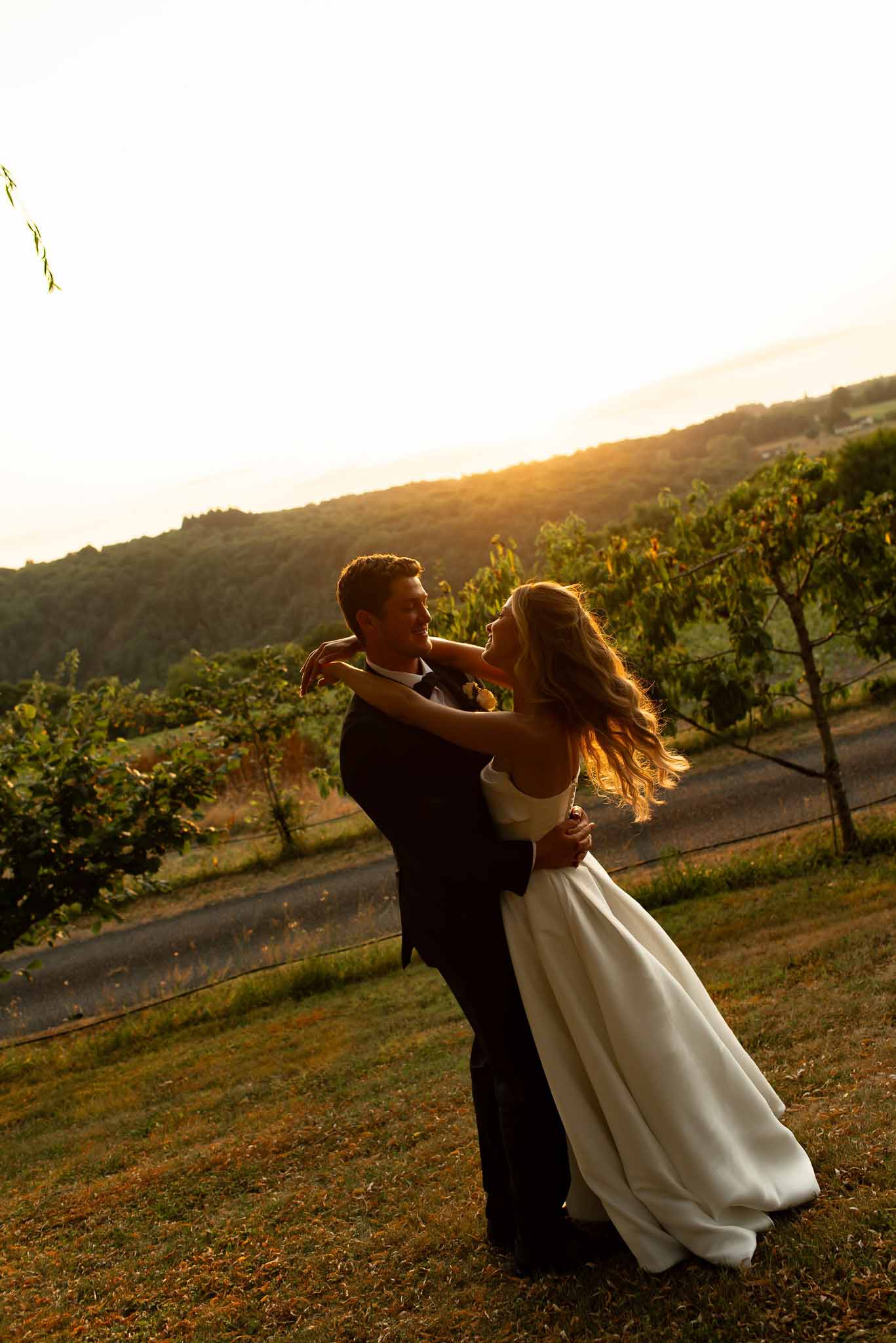 Couple dancing on grassy hillside overlooking vineyard rows at golden hour with warm backlit sunset