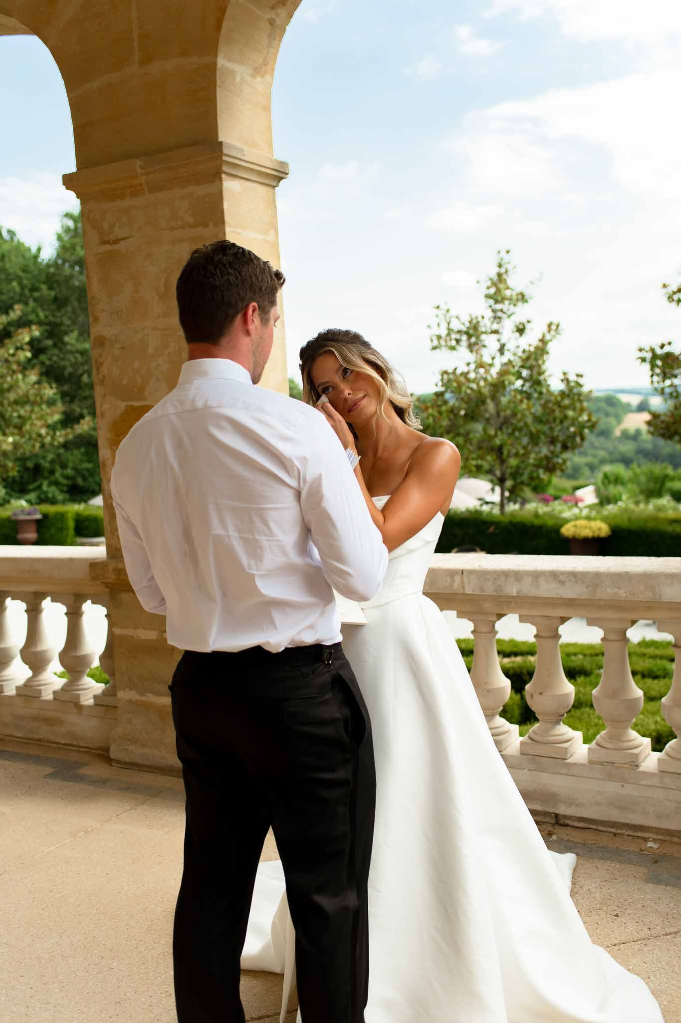 Bride wiping tear in ivory ballgown and white glove as groom holds her hand on chateau stone balustrade terrace