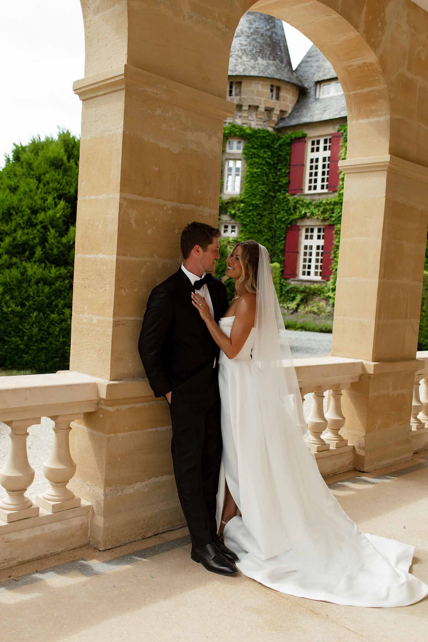Bride and groom embracing under a stone arch on a chateau terrace with ivy-covered facade and turret behind