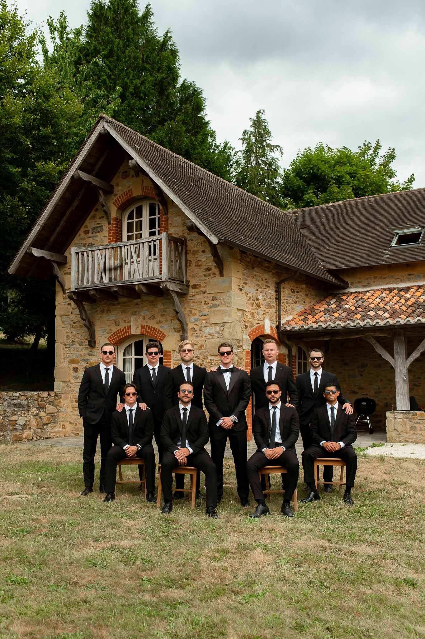 Ten groomsmen in black suits and sunglasses posed in two rows before stone farmhouse with balcony