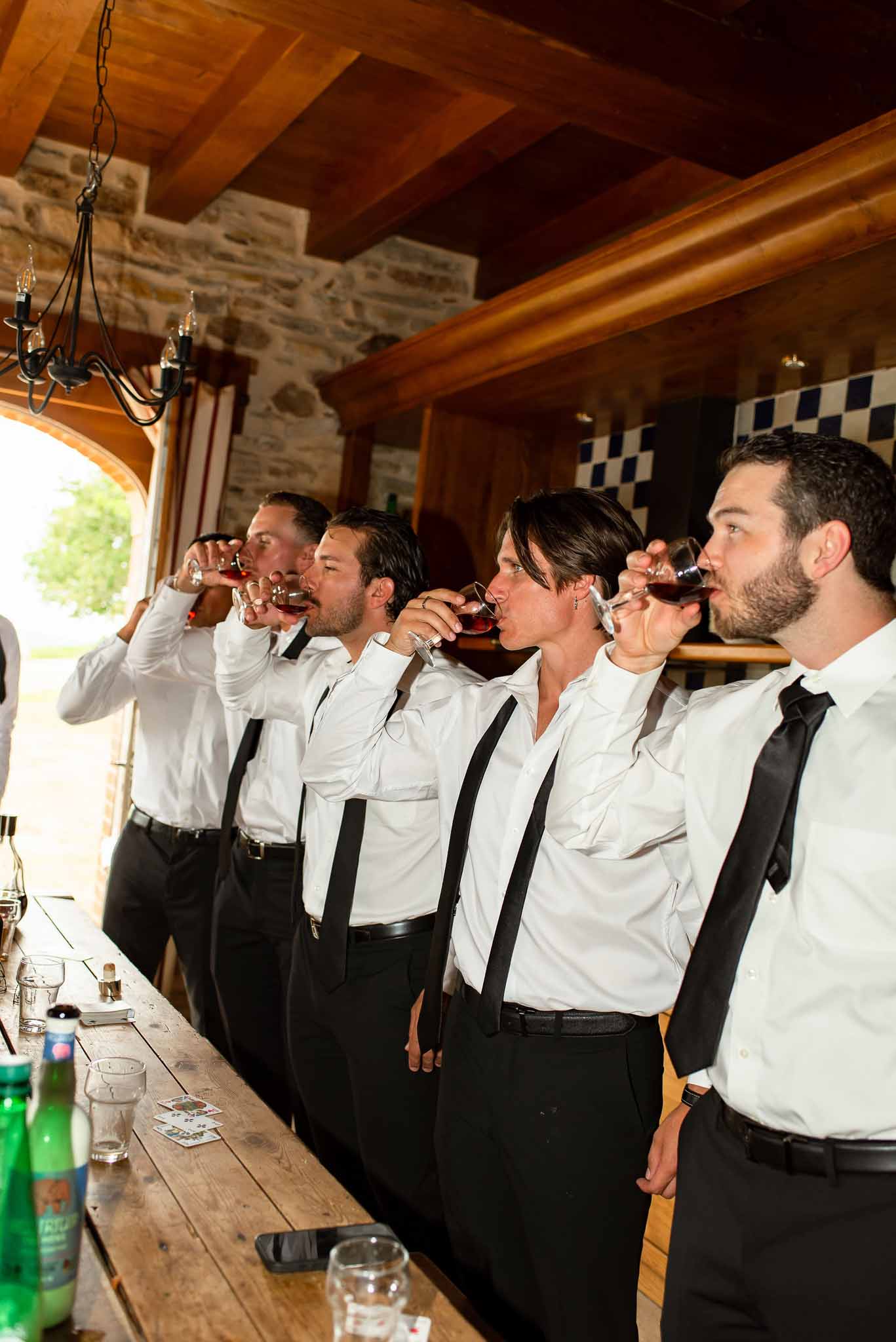 Four men in white shirts and suspenders drinking shots at rustic stone bar with wrought iron chandelier