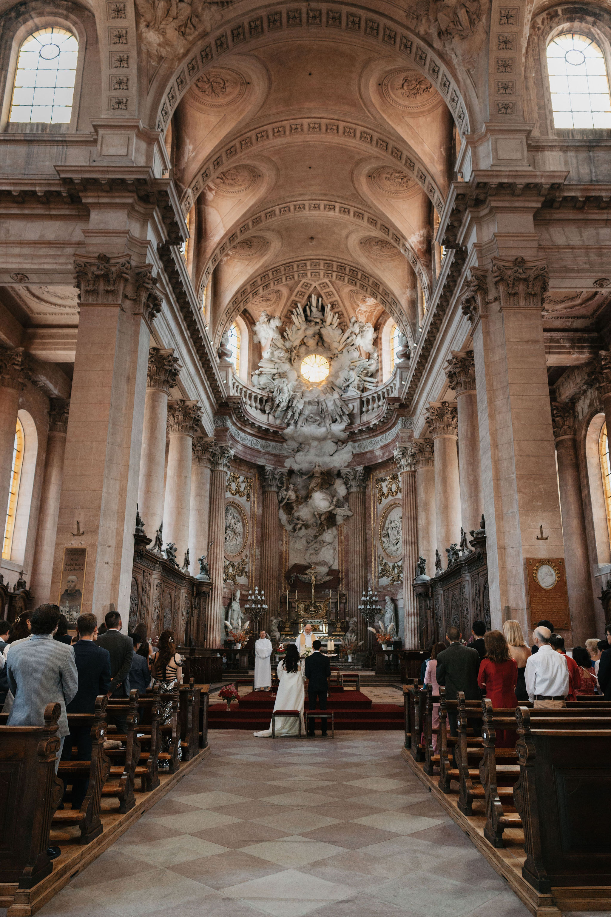 A religious wedding ceremony is taking place inside a grand Baroque-style church, shot from the rear of the nave looking toward the altar. The couple stands at the altar with what appears to be two officiants in white robes; the bride is wearing a full-length white gown with a train. Approximately 40–50 guests are seated in dark wooden pews on either side of the central aisle, dressed in formal attire including dark suits, a red dress, and a light grey suit. The church interior features vaulted coffered ceilings, tall ornate columns, red carpet on the altar steps, candelabras, and an elaborate sculptural altarpiece with white marble figures and gilded details at the far end. Small floral arrangements in warm orange and red tones are visible near the altar. The shot is a wide, symmetrical composition taken from a standing position at the back of the aisle. Potential venue feature image.