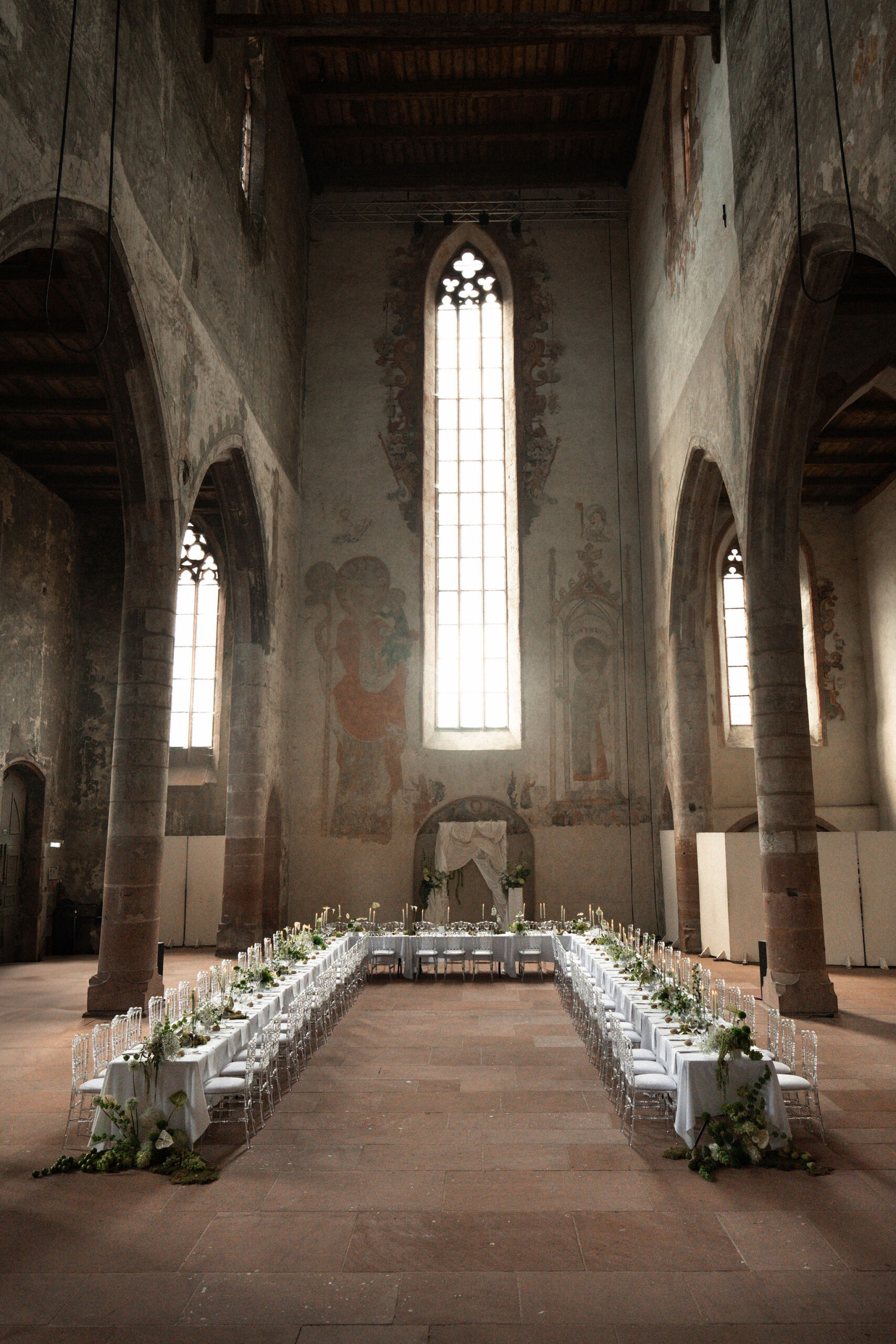 A wedding reception setup photographed from a wide, symmetrical perspective inside a converted medieval church or abbey with soaring stone walls, tall Gothic lancet windows, original frescoes, and a dark timber ceiling. Two long banquet tables run the length of the nave, dressed in white linen and lined with ghost chairs, meeting a head table positioned at the far end in front of a draped arch. The table runners feature trailing greenery and white florals, with taper candles in slim candleholders spaced along the length, and larger floral arrangements with green foliage and white blooms cascading to the floor at the table ends. The decor palette is white and green against the raw stone interior, creating a contrast between minimal modern styling and the aged architectural surroundings. No guests are present — this is a pre-reception setup shot. Potential venue feature image.