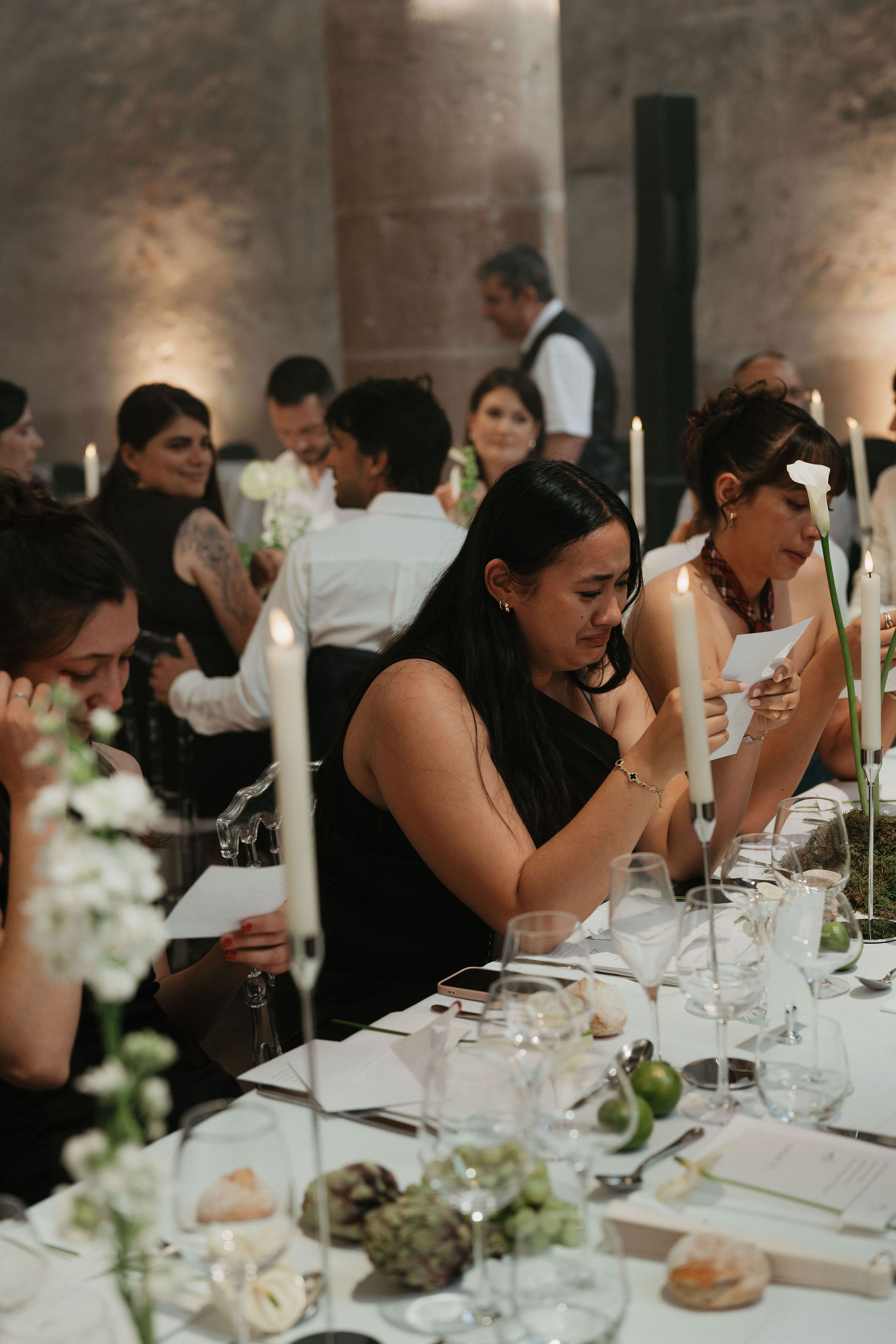 A wedding reception dinner is underway in a stone-walled indoor venue, likely a historic château or abbey, with warm uplighting against the textured stone columns in the background. Several guests seated at a long banquet table are visibly emotional, reading from small cards — one woman in a black one-shoulder dress appears to be crying, suggesting a speech or vow-reading moment is taking place. The tablescape features a white linen runner, tall ivory taper candles in thin holders, single white calla lilies, small clusters of white florals with greenery, artichokes, and green apples as decorative elements, with crystal glassware and formal place settings throughout. The overall decor palette is white, green, and natural, with a modern yet organic styling approach, captured in a candid medium shot from a slightly elevated angle along the table length.